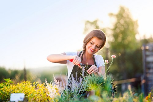 A smiling woman is tending to a garden in an outdoor space, using a small gardening tool to carefully prune or plant among a variety of flowering plants and shrubs. The garden features neatly maintained flower beds with colourful blooms and green foliage, bordered by paving slabs and a wooden fence in the background. The area includes lush, dense grass and a mixture of flowering and leafy greenery, with natural sunlight illuminating the scene and creating a warm, inviting atmosphere. The garden appears well-kept, reflecting active gardening maintenance typical in residential outdoor spaces within Battersea, London, with a focus on local landscaping and lawn care services provided by Garden Maintenance Battersea.