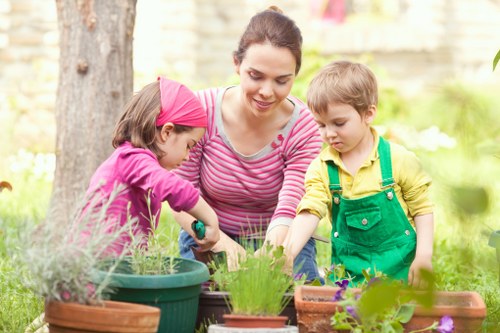 A woman with dark hair tied back, wearing a pink and white striped top, is engaged in gardening activities with two young children in a lush backyard. The girl, dressed in a pink long-sleeved top and pink headband, is holding a small gardening tool and appears to be planting or tending to a plant in a green pot. The boy, wearing a yellow shirt and green overalls, is observing closely, possibly assisting or learning. The garden features a variety of potted plants with different textures and shades of green, purple, and pink, arranged on a wooden surface. In the background, there are mature trees with textured bark, a stone or brick wall, and a well-maintained outdoor space with natural sunlight, suggesting mild weather. The scene captures a moment of outdoor gardening activity, suited to professional gardening and landscaping services, with a focus on family involvement and garden care in Battersea, London, with elements like soil, lush foliage, and plant containers visible in the outdoor environment.